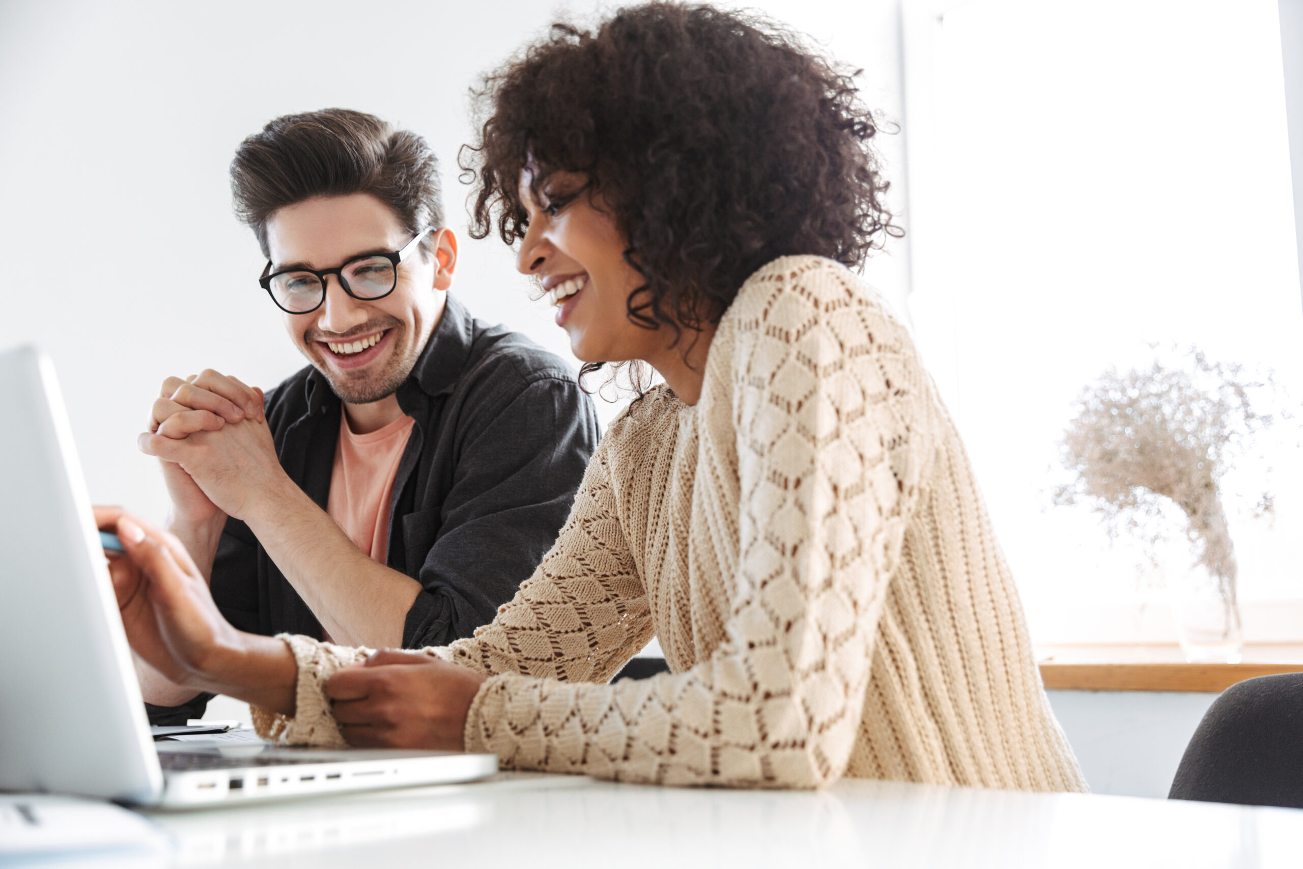 Cheerful young colleagues using laptop computer together while sitting by the table at office