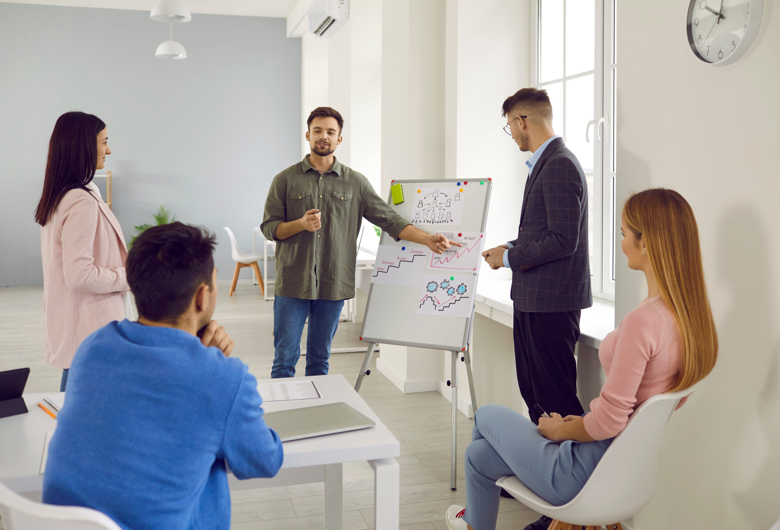 Young business team listens to ideas and suggestions of their male colleague during brainstorm in office. Young startup project manager standing in front of whiteboard with graphs speaks to colleagues
